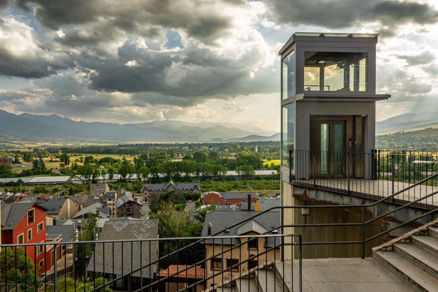 Ascenseur extérieur vitré accolé à un bâtiment, avec passerelle d'accès et vue sur une ville de montagne